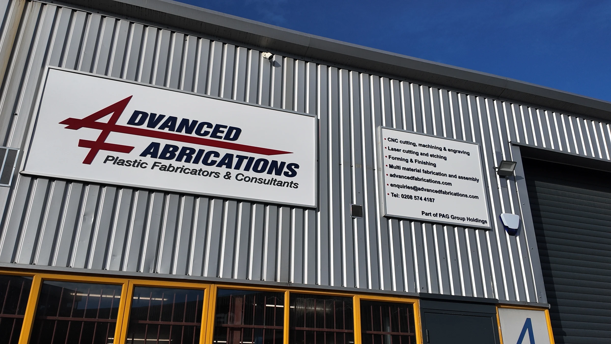 The front of the Advanced Fabrications unit from below: the sign, the services board and the roller door under a blue sky