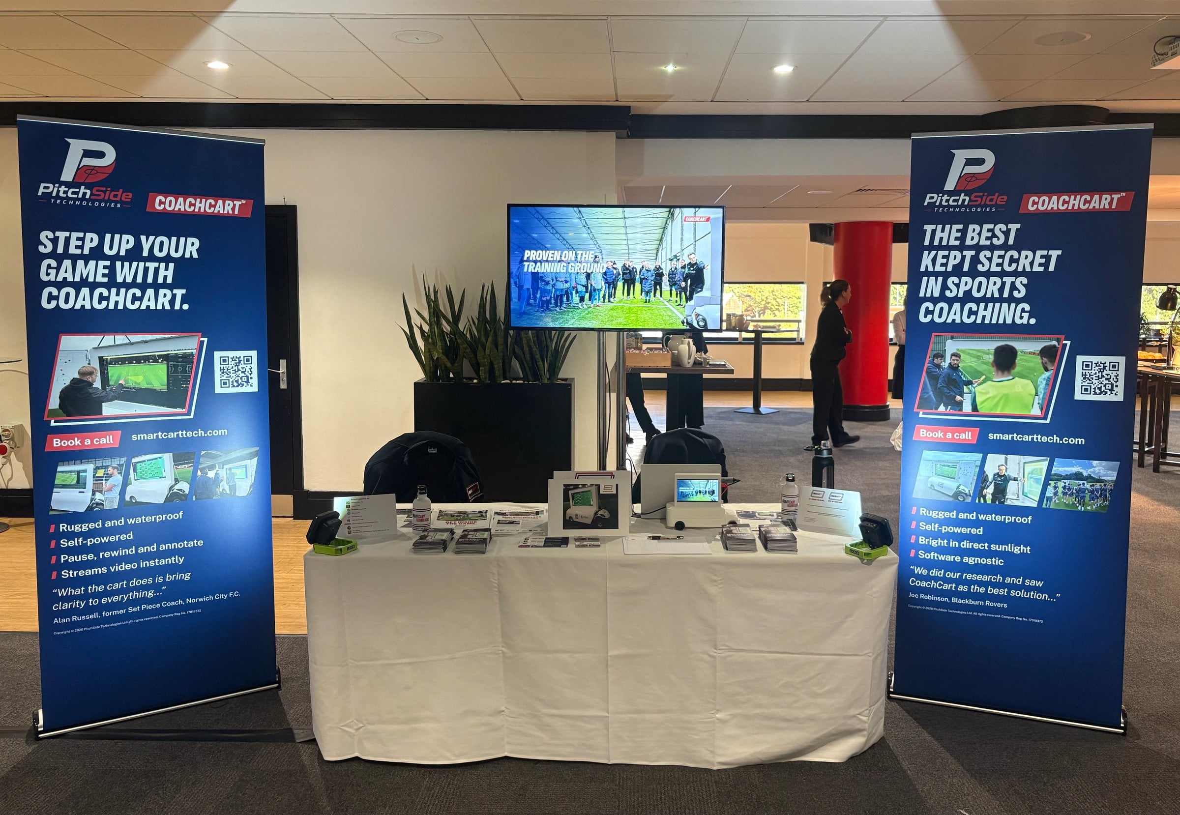 The PitchSide Technologies trade stand: two navy CoachCart pull-up banners either side of a screen and a table of printed leaflets and cards